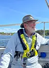Eldre mann med gul redningsvest sitter på seilbåt på rolig sjø i solskinn