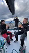 Tre personer på dekk på en seilbåt spiser mat på havet under grå himmel.