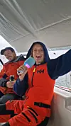 To personer i røde Helly Hansen-drakter sitter under et båttrekk, en holder en farget flaske.