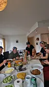 Fem personer rundt et bord med flere skåler, tallerkener og drikke i en lys stue.