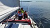 Seilbåt med hvitt seil, solcellepanel og to personer på dekk på stille vann.