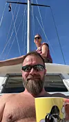 Mann med solbriller og skjegg sitter på en seilbåt med en gul kopp, med en kvinne ved masten bak.