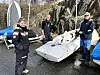 Tre personer ved flere små seilbåter på en våt brygge med fjell i bakgrunnen.