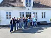 En gruppe mennesker står samlet foran et hvitt hus med rødt tak.