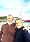 Et eldre par tar selfie ved en småbåthavn med seilbåter og brygge i bakgrunnen.