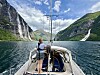 Seilbåt på fjord med bratte fjell, fossefall og en person ved masten.