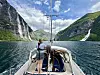 Seilbåt på fjord med bratte fjell, fossefall og en person ved masten.