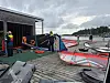 Personer klargjør seilbrett og seilutstyr på en brygge ved sjøen.