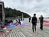 Personer står på en trepir med vannsportutstyr, brett og seil ved vannkanten under grå himmel.