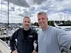 To menn står foran fortøyde båter ved en marina med master og overskyet himmel.