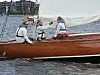 Tre personer sitter i en brun treseilbåt med hvitt seil på vannet nær en by.