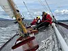 To personer i røde redningsvester om bord i en seilbåt med mast, tauverk og bølger rundt.