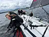 Mannskap på en seilbåt som krenger kraftig i bølger på åpent hav.