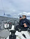 To personer om bord på en seilbåt med tauverk og rigg, med sjø og skyet himmel i bakgrunnen.