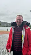 Smilende mann i rød jakke står ved en innsjø med grå himmel og landskap bak.