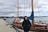 En kvinne står foran en seilbåt fortøyd ved en brygge ved vannet.