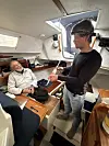 To menn i en liten seilbåt med treinteriør, én sitter og én står ved en luke.