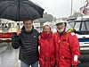 Tre personer står på en kai i regnvær med paraply, foran båter og en marina.