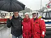 Tre personer står på en kai i regnvær med paraply, foran båter og en marina.