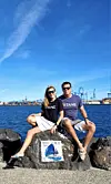 Et par sitter på steiner ved havnen med blå himmel, vann og kraner i bakgrunnen.