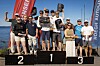 Flere personer står på en seierspall utendørs ved vannet, med nummer 1, 2 og 3 synlig.