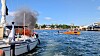 Flammer og svart røyk kommer fra en båt i en havn med andre båter i nærheten.