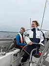 To personer står på dekk i en seilbåt med hav og grå himmel rundt seg.