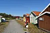 Grusvei med små hus og naust i et kystområde under blå himmel.