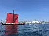 Vikinginspirert langskip med rødt seil og en hvit ferge på rolig sjø.