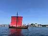 Tradisjonell båt med røde seil på vannet foran en kystby.