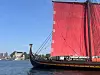 Et vikingeskip med rødt seil på sjøen nær en kystby.