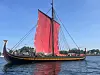 Tradisjonell trebåt med rødt seil på vannet nær land under blå himmel.