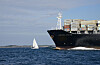 Stort containerskip og liten seilbåt på blått hav under en klar himmel.