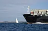Stort containerskip og liten seilbåt på blått hav under en klar himmel.
