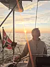 Seiler ved roret på en seilbåt med solnedgang over havet i bakgrunnen.
