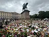 Blomsterdekket plass foran et monument med rytterstatue og mennesker under mørke skyer.