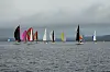Fargerike små seilbåter på grått fjordvann under mørke skyer.