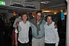 Tre menn smiler og poserer sammen i et innendørslokale.
