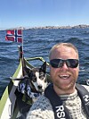 Smilende mann og hund i en liten båt på sjøen med norsk flagg og kyst i bakgrunnen.