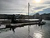 Trebåt med høye master ligger fortøyd i en byhavn i sollys.
