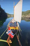 Seilbåt med norsk flagg og hvitt seil på stille vann mellom grønne fjell.