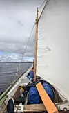 Utsikt fra dekket på en seilbåt mot masten, hvitt seil og åpent hav.