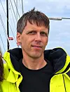 Mann i gul seiljakke om bord i en båt med master og tauverk i bakgrunnen.