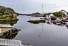 Liten kysthavn med båter, sjøhus, holmer og en rød bøye i stille vann.