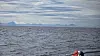 Åpent hav med fjell på horisonten og en person i en liten båt nederst til høyre.