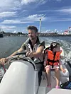 Voksen mann kjører motorbåt med barn i rød redningsvest foran seg.