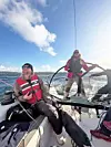 To seilere om bord i en seilbåt på havet med fjell og blå himmel i bakgrunnen.